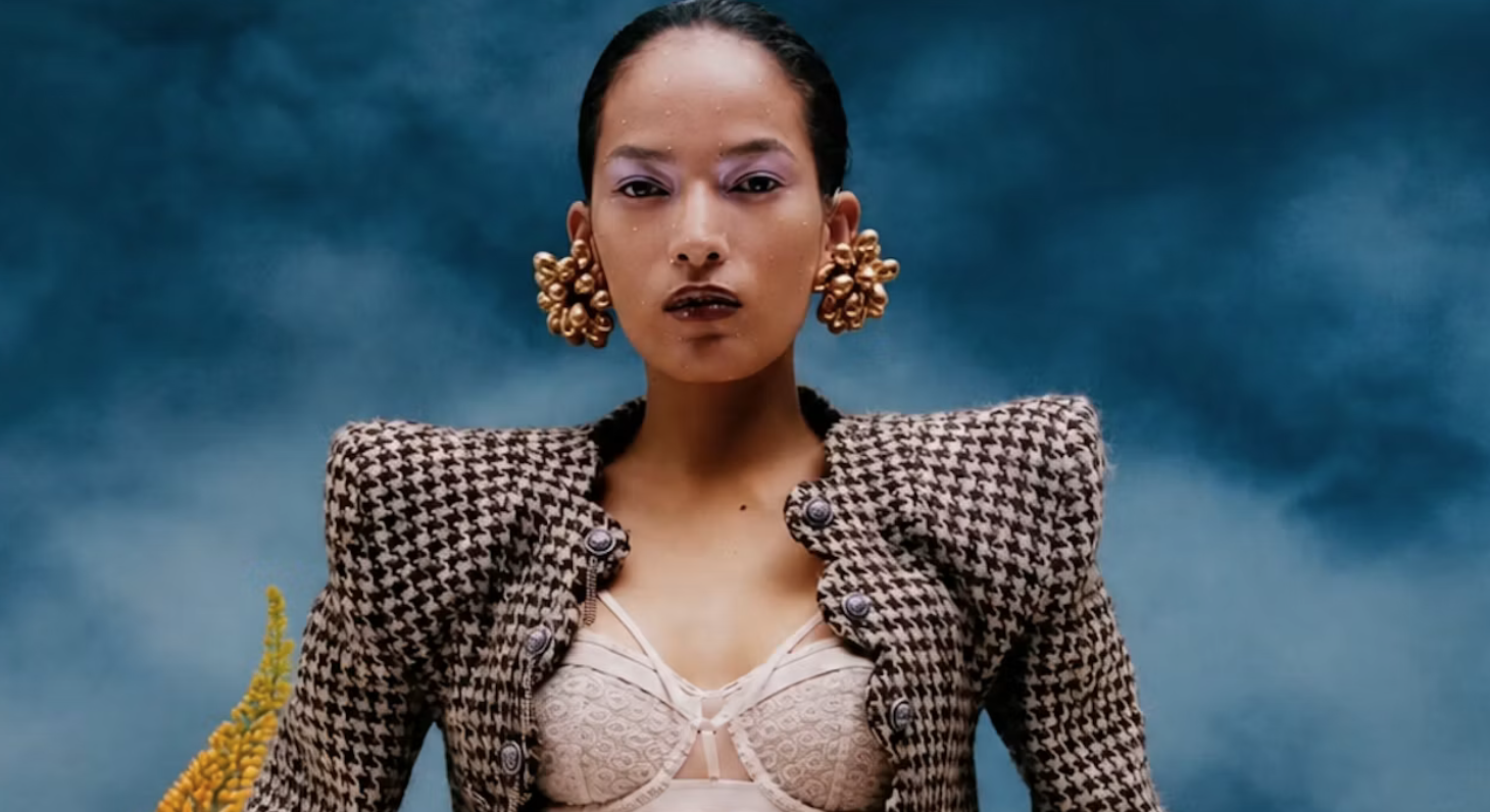 Fashion model wearing oversized sculptural gold earrings and a structured houndstooth jacket.